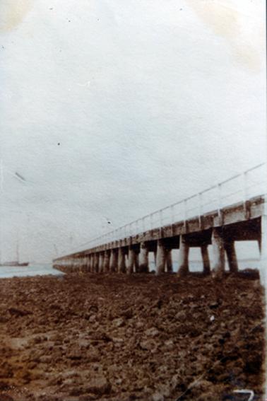 This image shows a long jetty jutting out into the water.  Rough, stony ground is in the foreground and water can be seen in the background.  All the jetty’s structure can be seen out of the water.  Railing runs along the left-hand side of the jetty which disappears out into the water.  A boat can be seen on the far left-hand side of the image.   