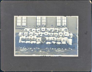 This image shows a group of thirty men, outside a building, posing for a photograph, in three rows.  There are ten men standing on a form in the back row, eleven men standing in the middle row and nine men sitting in the front row.  All men, except one, are in a white uniform of short sleeved t-shirts, pants to the knees, white shoes, black leggings and a black belt.  There is an emblem on the front of their shirts.  Fencing apparel is on the ground at the front.  The man in the centre of the front row is dressed in full military uniform and is holding a baton.  Three sets of windows can be seen in the wooden building behind the men.  
