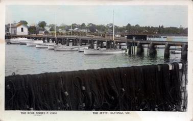 This image is of a jetty, taken from the end of the jetty, looking back towards the town.  There are at least eight fishing boats tied up to the jetty and more boats can be seen on the other side of the jetty and on the shoreline.  A boat-shed is on the jetty on the left-hand side of the postcard.  Fishing nets are hanging over a rail in the foreground.  Tall poles can be seen along the jetty.  Buildings and houses are in the background set amongst large tree.  Printed at the bottom of the postcard is: “The Rose Series P. 13936 copyright The Jetty, Hastings, Vic.”.