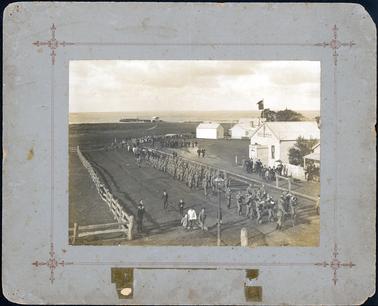 This image is of a military detachment marching in the street led by a military band.  A soldier on horseback is behind them and a team of bullocks, pulling a cannon on a gun carriage is following.  Men, women and children are bringing up the rear.  Wooden buildings with iron roofs, including the Library & Reading Room,  are on the right-hand side of the image.  A bay with jetty and fishing shed can be seen in the distance.  People are walking along either side of the parade.  A post and rail fence is seen on the left-hand side and a hitching post is on the right-hand side of the image.  A gas lamp is in the foreground.  The photograph is taken from an elevated height.  