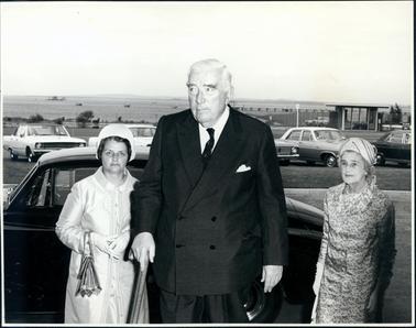 This image shows a man and two women standing in front of a car. The man is wearing a double breasted, dark coloured suit, white shirt and tie. He is holding a walking stick in his right hand. He has grey hair and dark bushy eyebrows. The woman on the left is wearing a light coloured coat, gloves, small hat and has a handbag over her right arm. Her hands are clasped at the front. The woman on the right is wearing a printed suit dress, gloves, hat and pearls. She is holding a handbag in her right hand. Five cars can be seen parked behind the group. Water, boats and a jetty can be seen in the distance, and land in the far distance. A small structure can be seen on the far right hand side of the image. The other two images show a group of people standing outside a brick building.  The large glass doors have BP on the handles.  