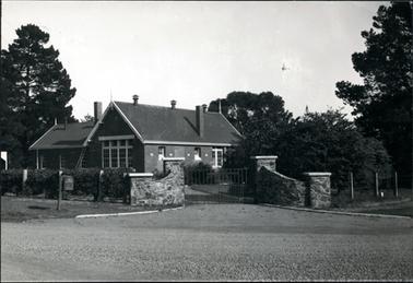 This image is of a brick school.  It shows the main building with four large windows at the front and a chimney on the right-hand side.  Three air vents are along the top of the roof.  An older building is on the left-hand side.  There is a curved stone entry with two wire gates.  A post and wire fence is on both sides of the gate.  A hedge is growing along the fence on the left-hand side.  A letterbox is on the footpath and the road is gravel.  Many large trees are beside and behind the buildings.  A ladder is leaning against the wall beside a window in the smaller building.  