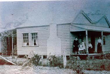 This image shows three women and two girls posing for a photo on the verandah of an old cottage with a double gable roof.  The two women on the right are dressed in long black skirts and long-sleeved white blouses.  The woman on the left is wearing a black long-sleeved dress with a white collar. The two girls are wearing black socks and white pinafores. The house has two windows and a brick chimney along the side.  Part of a rainwater tank can be seen underneath a lean-to at the back of the house.  A wire netting fence is across the front of the image and a large tree stump is in the foreground.  