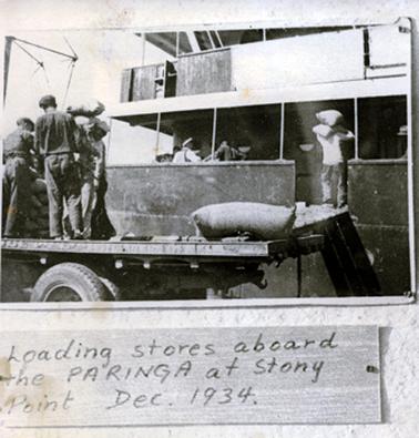This image shows a group of men loading supplies onto a boat.  There are several men on the deck of the boat and a man is carrying a bag on his shoulders from a truck to the boat.  In the foreground of the image the tray of a truck can be seen parked beside the boat, where four more men can be seen picking up bags from a big pile on the tray of the truck.  A large bag is lying on the end of the truck.  Printed at the bottom of the image is: “Loading stores aboard the PARINGA at Stony Point Dec. 1934.”.
