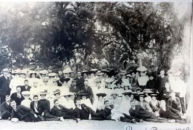 This image shows a large group of very well dressed men and women sitting and standing in front of large trees, posing for a photo. There are approximately sixty people.  All the women are wearing hats and the men are wearing three-piece suits.  Seven men are half lying on the grass in the front row.  They are leaning on their right elbows.  A man, third from the left is holding a baby on his lap.