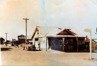 This image shows a street scene with shops along the right-hand side.  The building in the foreground is situated on a street corner and has three post verandah.  Large front windows are on either side of a door.  The words “Tea Rooms” are on the wall beside the window.  Two windows can be seen on the far right-hand side of the shop.  Advertising boards are leaning up against the building on the left-hand side.  A large high section of the building is behind the shop.  A utility is parked in the street.  Two tall power poles are beside the footpath on the left-hand side and a white street sign is on the far right-hand side.  
