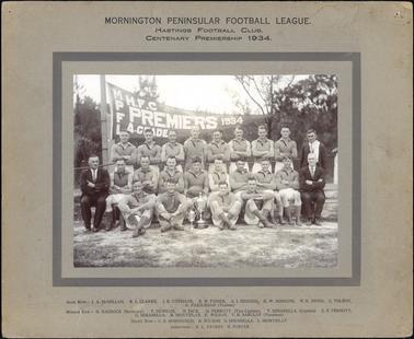 This image shows the Hastings Football club posing for a photograph when they won the Centenary Premiership in 1934 in the Mornington Peninsular Football League.  The image shows three rows of players along with their trainer, secretary and president.  The captain (middle row centre)  is holding a football and there are two trophies on the ground in front of him.  The Premiership flag is in the background.  Trees are in the far background and grass is in the foreground. All the players are in uniform.  The two men at either end of the middle row are wearing a suit and tie.  A man in the back row on the far right of the photograph is also wearing a suit and tie and has a towel across his shoulder.  Printed on the flag is: “H.F.C. PREMIERS 1934 A. GRADE” plus the initials: “MPFL”.  Printed on the mounting at the top is: “MORNINGTON PENINSULAR FOOTBALL LEAGUE. HASTINGS FOOTBALL CLUB. CENTENARY PREMIERSHIP 1934”.  All the names of the men are printed at the bottom of the mounting.  
