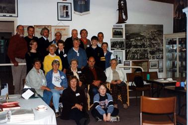 This image shows a group of twenty people, in four rows, posing for a photo.  There are seven men standing in the back row, six people standing in the second back row, five people sitting on chairs and two people sitting on the floor.  Tables and chairs are scattered around.  A trumpet and several photos and posters are handing on the wall behind the group.  Part of a glass display cabinet can be seen on the far right-hand side.  A cup and saucer sits on the table in the foreground.  