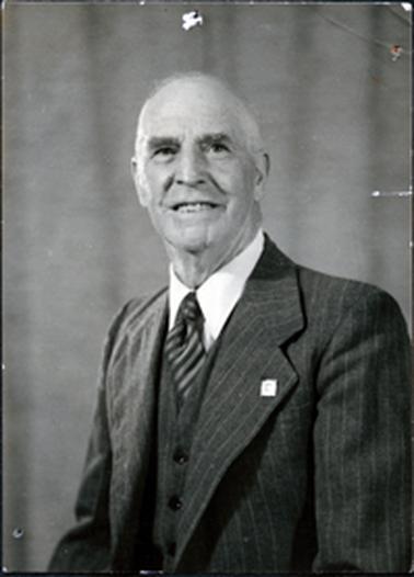 This image shows a portrait of a middle-aged man taken from the waist up.  He is wearing a three-piece suit with a striped tie and white shirt.  He has a badge on his left lapel.  He has a smile on his face, thick eyebrows and thinning hair.  