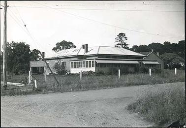 This image shows the Hastings & District Bush Nursing Hospital from the front, a single story brick building with a shaded verandah across the front facade and around part of the right hand side. A sign giving the name of the hospital is to the right of the building. A smaller building is on the left hand side and a gravel road is in the foreground, with a post and barbed wire fence in front of the building.