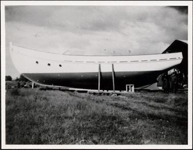 This image is of a boat on dry land which takes up the entire width of the photograph.  The top half of the boat is a light colour and the bottom half dark.  There are three round holes on the boat on the left-hand side of the photograph.  Two poles are standing up against the boat.  A group of men can be seen near a building on the right-hand side of the photograph.  A large expanse of grass is in the foreground and the sky is very cloudy.