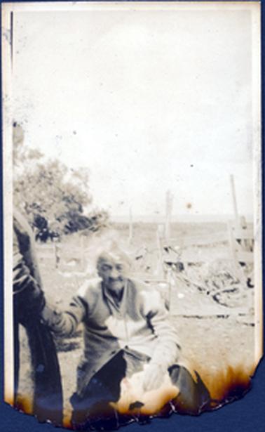 This image shows an elderly woman sitting outdoors holding a hand.  Only the arm of the other person can be seen.  She is wearing a dark skirt and a light coloured cardigan which is half buttoned up.  The photograph has been burnt and torn away at the bottom.  Part of an old post and rail fence can be seen behind the woman.  A paddock is in the background and a tree can be seen on the left-hand side.  