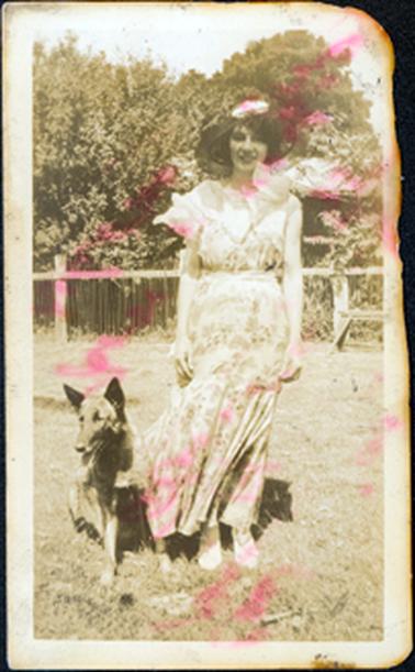This image shows a pretty young woman standing beside a dog, smiling for the camera.  She is wearing a wide-brimmed hat and a ankle length floral dress.  The large frilled collar and the gored skirt are blowing sideways in the wind.  Part of a house can be seen in the background amongst the trees and behind a fence. 