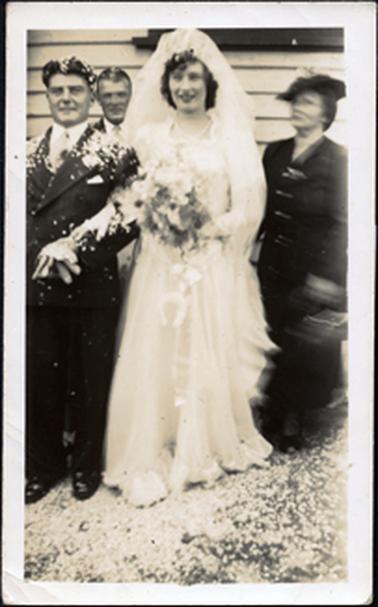 This image shows a bride and groom standing outside a building posing for a photograph.  A man’s head can be seen behind the couple and the blurred image of a woman wearing a dark coloured coat and hat, is on the right-hand side of the image.  The bride is wearing a high headpiece with a veil and is holding a large bouquet.  A horseshoe can be seen at the front of her shirt.  She has her right hand on the groom’s left arm.  A large amount of confetti is on the ground and on the groom’s suit.  