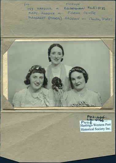 This image shows the top half of three beautifully dressed women posing for a photograph in evening/formal dresses .  They are all wearing necklaces, flowers on their dresses and flowers in their hair.  They are all smiling.  The photograph is mounted on cream embossed card and enclosed in a light brown embossed folder.  