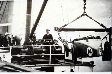 This image shows a racing car being loaded onto a boat.  The car has the number 14 in large numbers on the car.  There is a large crane lifting the car and a man, wearing a cap, is standing behind the crane engine.  Several people can be seen in the background watching.  