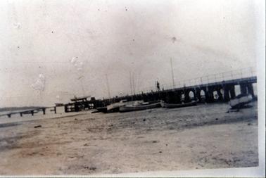 This image shows a pier with several boats standing at its side.  All the boats have tall masts.  A man can be seen standing at the centre of the pier.  Tall electricity poles can be seen along the pier.  A large expanse of water is in the foreground.  