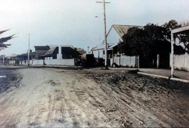This image shows a wide unmade road with buildings along the right-hand side.  Three power poles can be seen along the footpath.  A house with a white chimney is at the centre of the image.  A motor vehicle is parked on the footpath.  A white paling fence is in front of most of the buildings.  Verandah posts can be seen on the buildings at the end of the road.  A dog is sitting on the footpath near the first pole.  