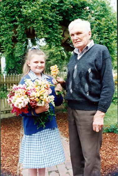 This image shows a young girl holding a bunch of flowers standing beside an elderly man who is handing her a single flower.  They are both smiling for the camera.  The girl is dressed in a blue check school uniform and is wearing a blue jumper.  Her hair is pulled back with a ribbon at the top.  The man is wearing brown pants and a pullover over his shirt.  He has grey hair.  A picket fence is behind them and they are standing on a brick path with bark chip on either side.  Some grass and part of a garden can be seen on the right-hand side and large trees are in the background.  