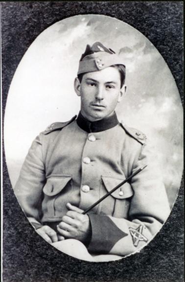 This image shows a young man in military uniform.  It is taken from the waist up and he appears to be sitting.  The image is in an oval shape.  His uniform has a high round dark coloured collar and three large buttons can be seen down the front of his jacket.  There are two large pockets with a smaller button showing at one.  There are epaulettes on his jacket, a dark coloured peaked cuff at his wrist and an insignia on the sleeve.   He is wearing a beret type hat with two buttons on the front band.  He is holding a small baton.  l 