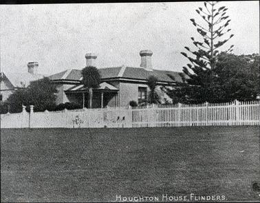 This image shows an impressive looking house with two very large chimneys on the roof.  It has a small verandah at the front, right-hand corner of the house.  There are stripes painted on the verandah roof and three verandah posts can be seen.  A large window is visible on the right-hand side of the image.  Trees and shrubs are at the front of the house and a large pine tree is on the right-hand side. A paling fence that includes a small front gate is across the front of the image.  It is unclear whether it is grass or a street that is in the foreground.  Part of another house, with a chimney can be seen on the left-hand side.  Printed across the bottom is: “Houghton House, Flinders The Cable Station Overlooking Flinders Pier”.