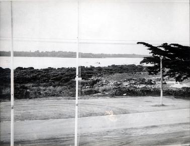 This image shows a foreshore lined with mangroves. A rubbish tip is on the right-hand side.  Large branches of a pine tree are partly visible beside the tip.  A stretch of water is across the middle of the image and land is in the background.  Several small boats dot the water.  Two small masts are in the foreground and a pole is on the right-hand side.  Wires run across the top  section of the photograph and a road is in the foreground.