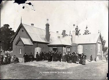 The image depicts the Hastings State School no. 1098 in 1913. The whole brick building is shown, with a timber entrance and a corrugated iron roof. There are about 30 adults and about 7 children grouped in front. There are 2 corrugated iron water tanks attached to the front of the building and what appears to be another at the back. At the bottom of the image is written in white “Re-opening S.S. 1098 Hastings, S:6.7.13”