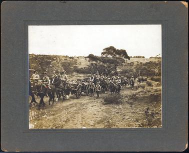 This image shows a large group of over thirty military personnel travelling through the countryside.  They are all in full military uniform and most of the soldiers are on horseback.  Three horse-drawn wagons can be seen.  There is a large tree behind the men in the centre of the image.  Other large trees can be seen on the far right-hand side of the photograph and fence posts can be seen in front of these trees.  The hill side in the distance is dotted with trees.  