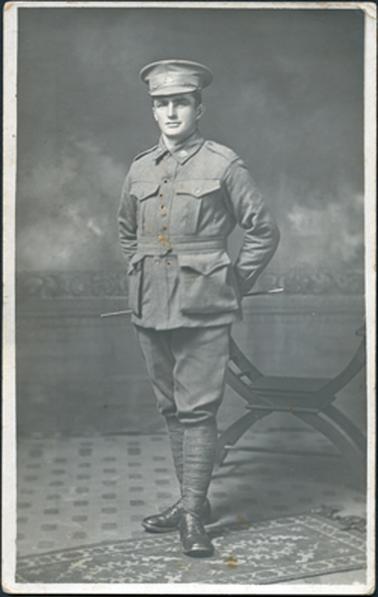 This is a full-length portrait of a soldier in full uniform standing with his hands behind his back, posing for a photograph.  There is a studio backdrop on the wall behind him and he is standing on a carpet.  A chair is on the right-hand side of the image behind the soldier.  