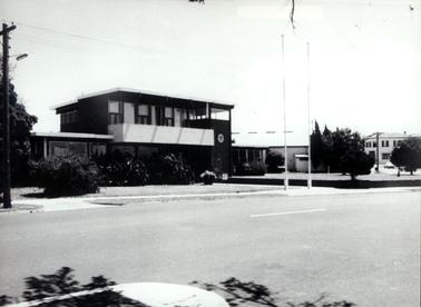 This image shows a building on two levels, with a wide street in the foreground.  There is a low section on each side of the building.  The two storey section in the middle has a verandah across the front.  Windows and doors are across the front of both sections. The building has a flat roof.  Several shrubs are across the front of the building.  Two tall flag poles are on the footpath at the front.  Another two storey building is on the far right-hand side.  A wide road is in the foreground.  A tree and electricity pole are on the footpath on the far left-hand side of the image.  