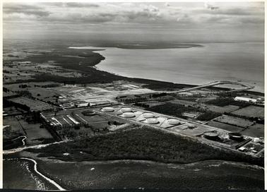This image shows a large industrial site with eight large tanks.  A large section of prepared ground is at the centre of the image.  A road leads out onto a circular wharf in the water.  Water and a shoreline are at the top left-hand section of the image.  Paddocks, trees and a creek can be seen.  