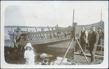 This image shows a group of men beside a partly built boat who have stopped work to pose for a photograph.  A little girl, dressed in white and wearing a large white hat is in the foreground.  The boat is helf up by two poles at the front.  There are seven men in the photograph.  The man on the left is wearing a waistcoat, hat and his long sleeves are rolled up.  There are two men wearing caps behind the boat who are only partly visible.  On the right-hand side of the photograph are a group of four men.  The one on the left is wearing a waistcoat, white long sleeved shirt rolled up and is holding a hat in one hand and a saw in the other.  Behind these men is a sail and mast.  There is water and land in the background. 