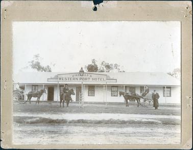 This image is of a single storey, bungalow style building with three small doors and six windows across the front.  A large board on a stand at the front reads: “O’Reilly’s Western Port Hotel”.  There is a horse and cart with a man driving on the left-hand side of the photograph.  Another man on horseback is near the hotel sign and on the right of the photograph is another horse and cart with a man in the cart and another man standing beside the cart wearing a three piece suit, hat and overcoat.  A gaslight is above the sign and a ladder is under the sign.   Large trees can be seen at the back of the building.  A road is in the foreground. 