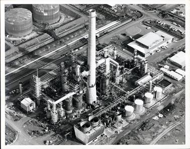 This image shows an aerial photograph of an oil refinery.  There is a very large chimney in the centre of the image and different size tanks can be seen at the top and bottom of the image.  There are several building on the right-hand side of the image and several cars can be seen both in front of the buildings and in a car park.  