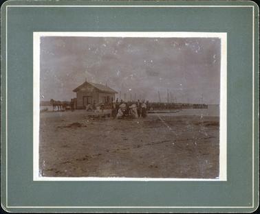 This image shows a group of people on the sand, posing for a photo.  There are seven people standing and three sitting in front of them.  Everyone is well dressed and the ladies are wearing large hats.  A fishing shed and jetty is behind the group.  Another group of people are in front of the shed and a horse and cart are standing in front of the shed.  Many fishing boats can be seen beside the jetty and fishing nets are hanging over the jetty rail.  Land can be seen in the distance and some water with a large stretch of sand is in the foreground.  