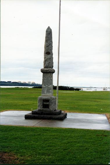 This image shows a War Memorial mounted on a two step concrete base and sitting on a large concrete slab.  A white flagpole is to the right.  Writing and plaques can be seen on the monument.  Water is in the background, dotted with boats.  Large tanks can be seen behind trees on the far left-hand side of the image.  Green lawn is in front and behind the monument.  