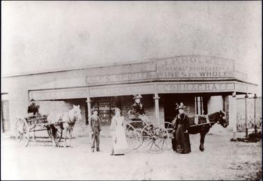 This image is of a General Store with a group of people on the roadside at the front of the store.  On the left-hand side of the image is a man in a two wheel spring cart with a light coloured horse.  He is wearing a hat and has a dark beard.  The other vehicle is a plaeton being pulled by a dark horse with a lady is sitting in the seat.  She is wearing a large hat and a dark coat.  Standing beside the plaeton is a lady in a dark dress, large hat and holding an umbrella.  A lady in a light coloured dress with a white apron is standing beside the back wheel of the plaeton.  Behind her is a man, wearing a three piece suit, hat and tie.  They are all looking at the camera.  The shop is of brick construction with a verandah on two sides with wooden posts on the footpath.  The signs on the shop are: “J.P. Holmes General Storekeeper Ales, Spirits, Wines etc. Wholes Bakery Grocery Corn & Chaff.  The shop is currently operated by Richies, IGA Supermarket.