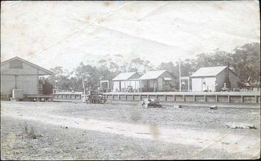 This image shows the Hastings railway station, looking towards the east. Behind the platform are 3 small buildings, one with a verandah which was presumably the waiting room. There are 2 signs with the word “HASTINGS” on them. Four people are on the platform and there are 2 horses and buggies drawn up behind, with goods piled up on the platform. To the left on the other side of the tracks is a goods shed. In the foreground is a pile of fish boxes marked Borrett, one of the fish salesmen at the Melbourne Fish Market. A pile of fruit boxes is also in the foreground.