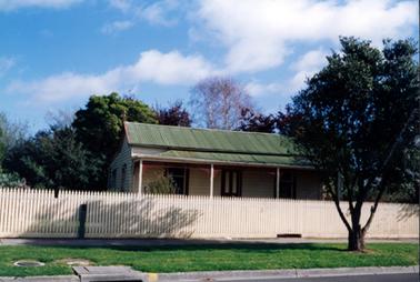 This photograph shows a small cottage taken from the street.  It has a green galvanized iron roof and a four post verandah.  A door and two windows are at the front and two windows can be seen down the left-hand side of the cottage.  A cream coloured picket fence is across the entire front of the image.  A concrete footpath, green grass with a tree and a gutter and part of a street are in the foreground.  Large shrubs and trees are to the left of the cottage.  A large tree with no leaves is behind the cottage.  