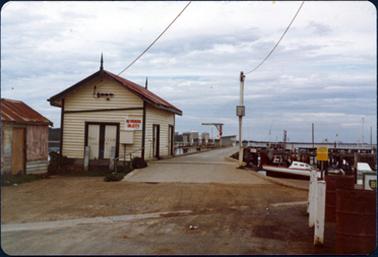 This image shows a fishing shed and a pier with part a small shed on the left-hand side.  Both the buildings have rusty roofs.  The sign ‘no parking on jetty’ is on the wall of the larger shed.  Boats can be seen in the water on the right-hand side of the image along with several tall masts.  Large white tanks can be seen in the far background.  Two white posts beside two rusty 44 gallon drums are in the foreground on the far right-hand side.  Part of a BP sign can be seen.  A gutter runs across the dirt road in the foreground.  Water and land can be seen in the far distance.