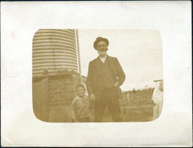 This is a faded image of a man and a small boy standing in front of a galvanised iron water tank on a brick stand.  The man is wearing a three-piece suit and a hat.  He has no tie, but his shirt is done up to the top.  He has a pipe in his mouth and a short white beard. His left hand is in his trouser pocket and his right hand is holding the hand of a small boy.  The boy is wearing a jacket with a rounded collar and buttons down the front.  There is a fence in the background and trees in the far distance.  An unidentified ‘object’ is on the right-hand side of the photograph.  