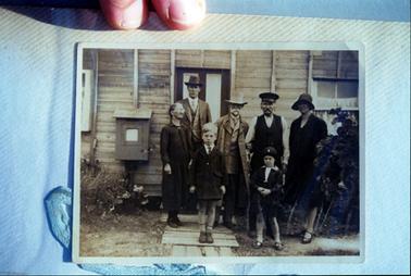 This image shows a group of three men, two women and two children standing outside a weatherboard house, posing for a photo.  Both women are dressed in black.  The woman on the left is tiny and wearing spectacles.  The other woman is tall and is wearing a hat.  The two men on the left are wearing three piece suits, hats and coats.  The other man is wearing a uniform.  The little boy and girl are wearing dark coats above the knee and shoes and socks.  A panelled door is behind the group and windows can be seen on either side.  A letter box is attached to the wall on the left-hand side of the building.  Shrubs are on either side of the group.  