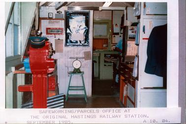 This image shows an office in the foreground with a doorway leading into another office.  There is a large red machine on the left-hand side of the room.  A set of scales are sitting on a stand in the centre of the image and a large picture is on the wall above it.  Written across the bottom of the picture is: “go relaxed by rail”. Other objects and papers on clip boards are around the walls.  Drawers and cupboards are on the right-hand side of the door way and part of a jacket can be seen hanging on the wall in the foreground, on the right-hand side. The view into the next room shows the right arm and shoulder of a person.  Cupboards a papers clipped on the wall are in the background.  Printed across the bottom of the photograph is: “Safeworking/parcels office at the original Hastings Railway Station. September 1985. A10. 04”.