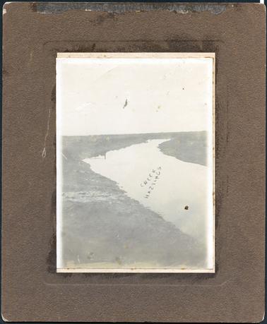 This image shows a creek with a shore line on either side.  There is some scrub on the banks of the creek and trees on the distant horizon.  The image is very faded.  Printed in the middle of the image is: “Creek Hastings”.  The letter ‘s’ in Hastings has been printed back-to-front.   
