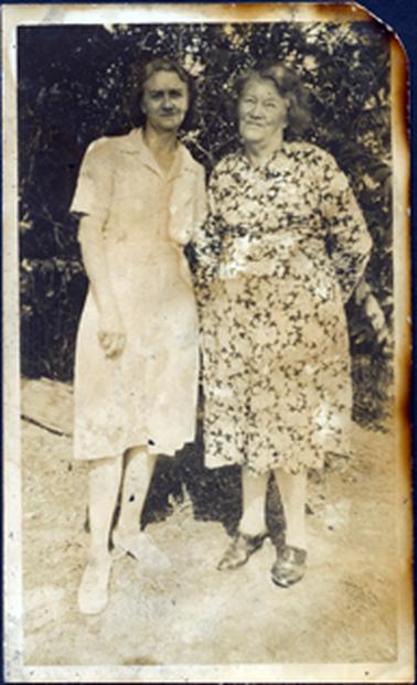 This image shows two women standing together in front of a garden, posing for a photo.  The woman on the left is wearing a light coloured, short sleeved dress.  The other woman is older and wearing a long sleeved, floral dress.  Garden and shrubs can be seen behind them.  