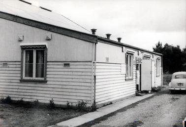 This image shows a woman standing in the doorway of a small weatherboard building, looking at the camera.  Only part of the building is visible.  One window is on the left of the building and three windows can be seen down the side of the building.  A sign above the door reads: “Shire Engineer Shire Secretary”.  Another sign is on the wall beside a window.  Three air vents are on the roof.  Large trees are in the background on the right-hand side of the image and small plants are across the front of the building.  A sedan motor car is parked on the driveway on the right-hand side of the building.  Its number plate is “RZ 827”.  