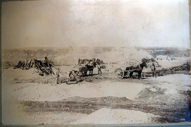 This image shows four men with two horses and carts, working on a road.  Two of the men are standing beside a horse and cart.  Two men in the foreground appear to be filling large sack bags.  Piles of stone and timber are behind the group and the buildings and trees of a town can be seen in the far background.  A rough gravel road is in the foreground.  