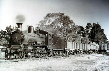 This image shows a goods train with a steam engine.  There are five goods wagons with a guard’s van at the rear.  Steam can be seen around the top of the engine.  Grass is in the foreground and tall trees are behind the train.  Part of a fence and building can be seen on the far left-hand side of the image.  