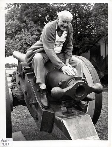This image shows a very happy grey haired man sitting on the top of a gun barrel smiling for the camera.  He is holding a rag in both hands and leaning forward polishing the barrel.  He is dressed in light coloured trousers, a white T-shirt with the words ‘Western Port Festival’ on the front and a open cardigan. The gun is sitting on a field carriage on the lawn in front of a building.  There is a large tree in front of the building and a tree fern on the right-hand side in the background.  Part of a car can be seen in the background on the left-hand side and a line of trees in the distance.