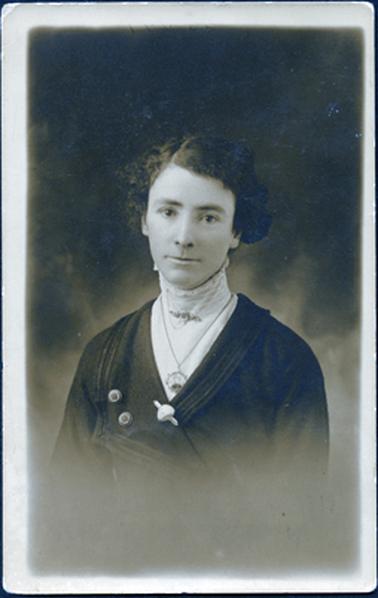 This image shows a head and shoulders portrait of a well dressed young woman.  She has a white blouse with a very high collar covering her neck with lace at the top. She has a brooch with a safety chain pinned at the neck and a chain and pendant, showing an image, is around her neck.  Her dark dress/jacket has a small collar on one side and a large collar on the other side with two buttons and a brooch.  He curly hair is neatly combed and parted on the right-hand side.  